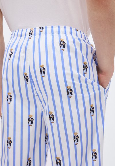Παντελόνι με μπλε και λευκές ρίγες, διακοσμημένο με μικρές αρκουδίτσες που φορούν ναυτικό μπλέ σακάκι και κρατούν φλιτζάνια, φορεμένο από ένα άτομο με το χέρι στην τσέπη.