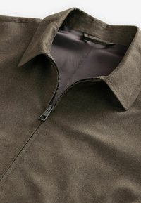 Braune Jacke mit Reißverschluss, die einen Kragen hat, einen glatten Stoff und ein metallisches Reißverschlussdetail aufweist. Das Innere ist in einem kontrastierenden Grau gehalten.