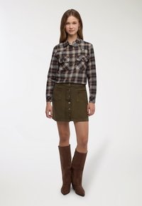 Camicia di flanella a quadri marrone e crema, con due tasche sul petto. Minigonna verde oliva con bottoni, finitura testurizzata e due tasche frontali. Stivali marroni fino al ginocchio.