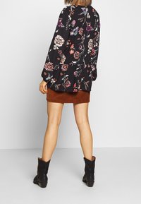 Schwarze Bluse mit langen Ärmeln und floralem Muster, die farbenfrohe Blumen und einen Peplum-Saum aufweist. Kombiniert mit einem braunen Cordrock und schwarzen Stiefeletten.