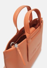 Borsa in pelle color tan di Calvin Klein con doppie maniglie, patch con logo esterno e chiusura a zip superiore, mostrata in primo piano angolato.