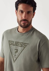 Olīvu zaļš kokvilnas T-kreklu ar lielu trīsstūrveida grafiku, kurā redzams "GUESS" logo un teksts, noapaļota apkakle un īsas rokas. 