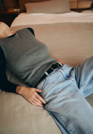 Top gri fără mâneci și blugi din denim de un albastru deschis, cu o curea neagră. Țesătura pare moale, iar decorul include o cuvertură de pat în tonuri neutre.