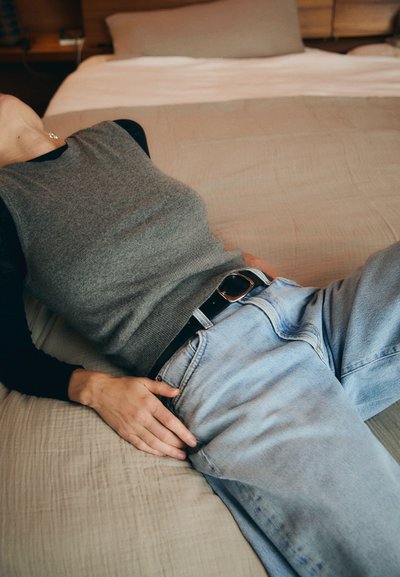 Un haut sans manches gris associé à un jean en denim bleu clair avec une ceinture noire. Le tissu semble doux et le décor présente une couette aux teintes neutres.