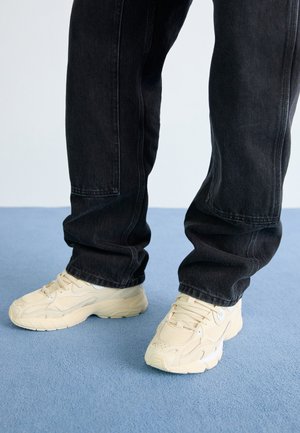 Personne portant un jean noir et des baskets épaisses crème debout sur un sol recouvert d'un tapis bleu.