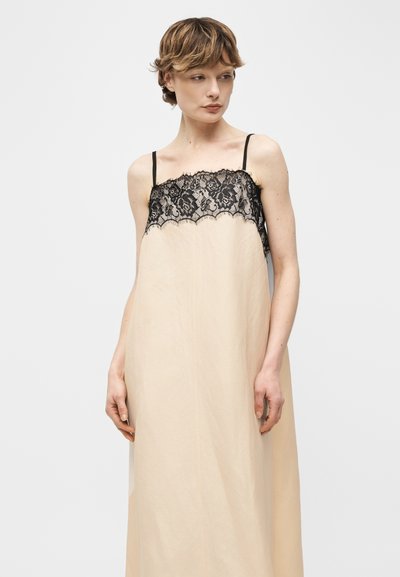 Νεαρό άτομο με κοντά μαλλιά φοράει μπεζ φόρεμα χωρίς μανίκια με μαύρη δαντέλα, στέκεται μπροστά σε απλό ανοιχτό γκρι φόντο.