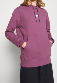 Lila hoodie i mjukt tyg. Har en framficka, ribbade ärmslut och en liten logopatch på bröstet.