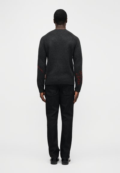 Grå sweater med tekstureret strik, med røde bogstaver på ærmerne. Kombineret med sorte jeans og sorte sko, vist bagfra.