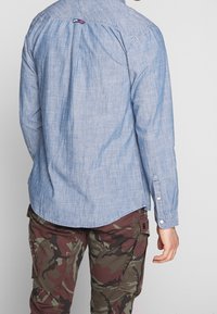 Camicia button-up in chambray blu chiaro, maniche lunghe, orlo curvo, con un piccolo etichetta rosso, bianco e blu sul retro vicino al colletto. Design casual.