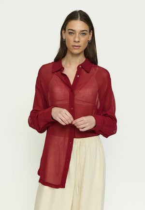 Pruunide pikkade juustega naine kannab läbipaistvat punast nööpidega pluusi koos sobiva rinnahoidja ning beežide paelaga pükstega, seistes ühevärvilise tausta ees.