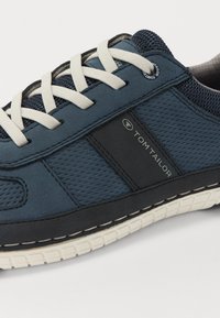 Baskets de sport bleues avec une tige en mesh texturé et en cuir, lacets crème et semelle extérieure en caoutchouc blanche. Présente un accent de logo noir.