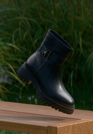 Bottine noire en cuir avec détail de boucle flottant au-dessus d'une surface en bois, sur un fond flou d'herbe verte.
