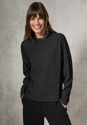 Schwarzes Langarmoberteil mit einem wellenförmigen weißen Muster, runder Ausschnitt, lockere Passform, kombiniert mit schwarzen Hosen. Glatte Textur, lässiges Design.