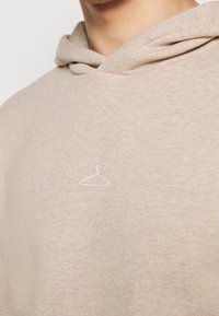 Sudadera con capucha beige hecha de tela suave, con un pequeño logo de una percha blanca bordado en el área del pecho.