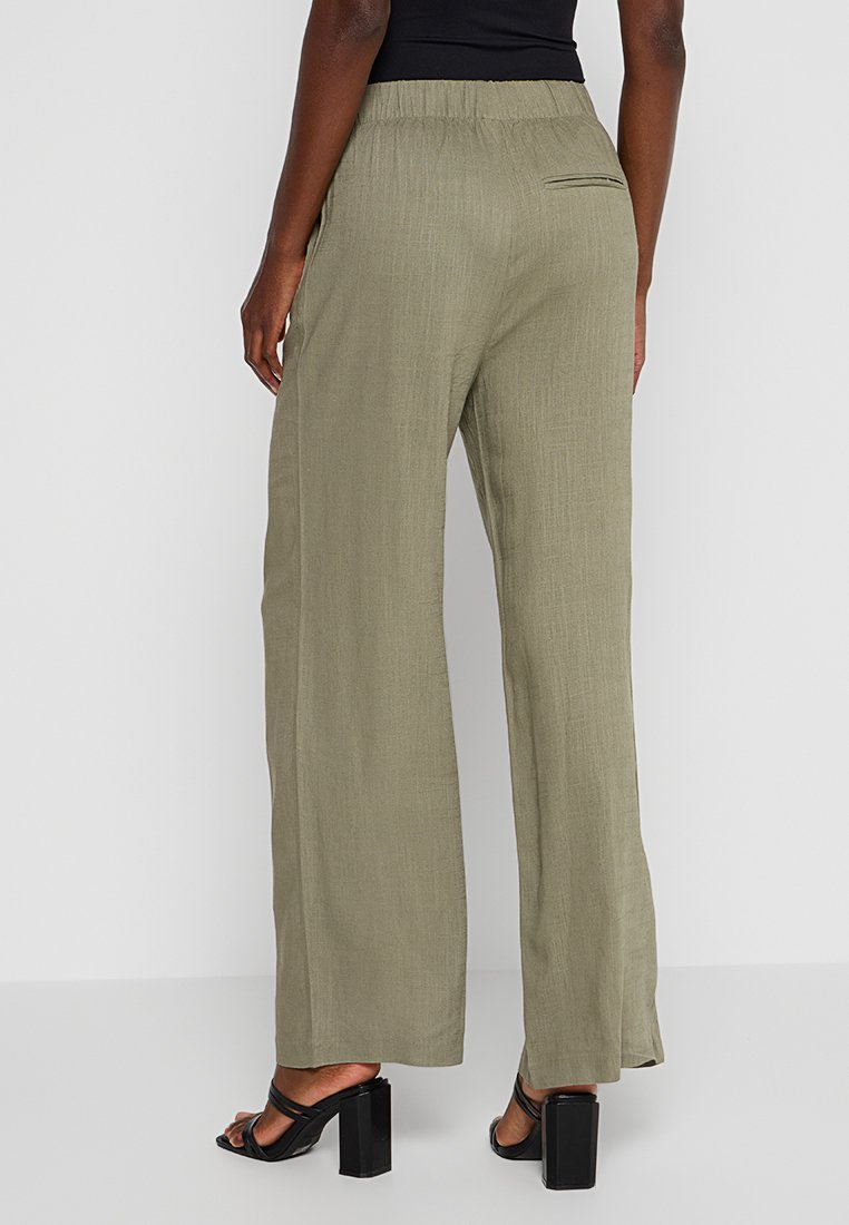 Olivgrüne, weit geschnittene Hose aus einem strukturierten Stoff mit elastischem Bund und einer Gesäßtasche. Kombiniert mit schwarzen Absatzsandalen.