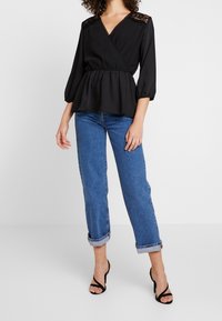 Black wrap blouse with lace detailing on shoulders, balloon sleeves, and peplum waist. Paired with rolled-cuff blue denim and black heels.