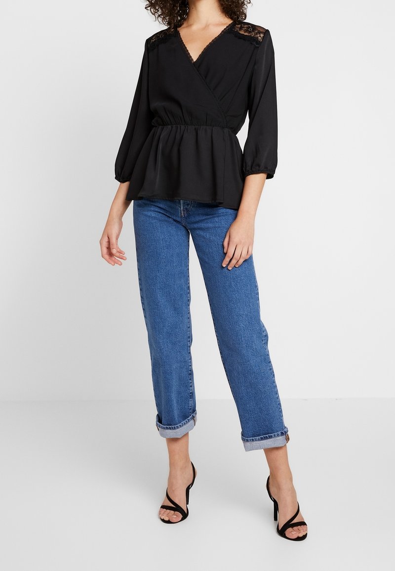 Black wrap blouse with lace detailing on shoulders, balloon sleeves, and peplum waist. Paired with rolled-cuff blue denim and black heels.