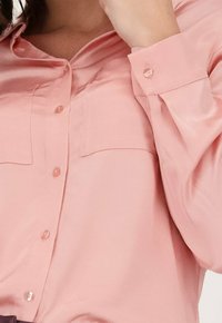 Lichtroze satijnen overhemd met knopen, voorzien van twee borstzakken en met knoopmanchetten, met een gladde textuur en een getailleerd ontwerp.