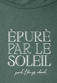 Zelené bavlněné tričko s výrazným bílým textem: "ÉPURÉ PAR LE SOLEIL" nad menším textem "dobré věci přicházejí." Hladká textura, kulatý výstřih.
