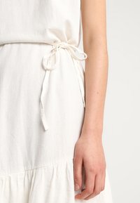 Vit linnklänning med dragsko vid midjan, utsvängd nederdel och slät yta. Tyget verkar lätt och luftigt.