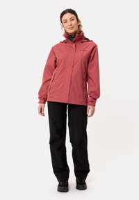 Rode waterdichte jas met verstelbare capuchon, ritszakken en elastische manchetten, gecombineerd met zwarte broek en zwarte schoenen.