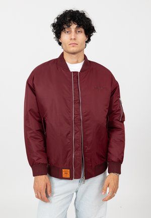 Bordowa kurtka typu bomber z przednim zamkiem błyskawicznym, ściągaczami na mankietach oraz pomarańczową naszywką. Posiada kieszeń na zamku na lewym rękawie.