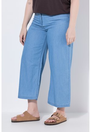 Person iført lyseblå vidde jeans og beige sandaler med spænder, der står mod en ensartet lys baggrund.