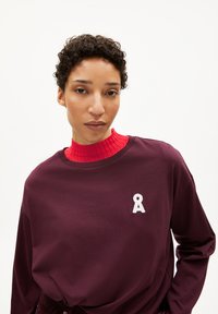 Bordeaux lange mouwen shirt met een ronde hals en een witte geborduurde lintlogo op de borst, gedragen over een geribbeld rood coltrui.