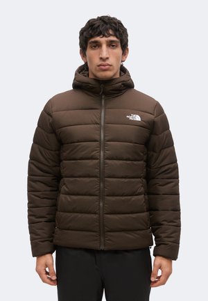 Man draagt een bruine doorgestikte North Face jas met capuchon, staand met het gezicht naar voren tegen een effen achtergrond.