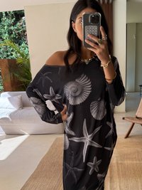 Vestido preto oversized com padrões de conchas brancas e estrelas do mar. Tecido macio e leve; design à mostra dos ombros; acessórios minimalistas e fundo texturizado.