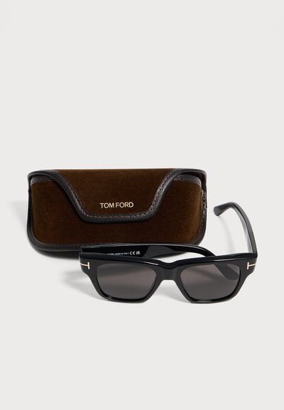 Lunettes de soleil rectangulaires noires Tom Ford avec verres foncés à côté d’un étui Tom Ford en daim et cuir marron sur fond clair.