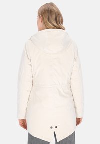 Kvinde iført en lang, off-white hættetrøje vinterjakke set fra ryggen, med samlet talje og et lille slidsdetalje i kanten.