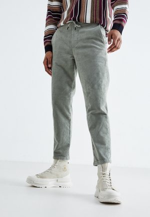 Man draagt lichtgrijze corduroy broek, beige hoge sneakers en een meerkleurige gestreepte trui, staand op een witte vloer en tegen een witte achtergrond.