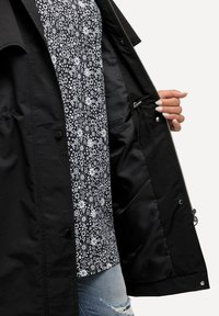 Musta takki vetoketjulla, jossa on kukkakuosiset vuori. Ulkopinta on sileä, ja siinä on näkyvää metalliosaa ja taskuja.