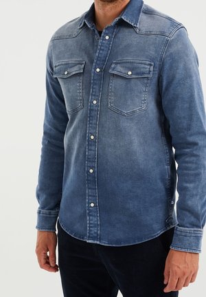 Homme portant une chemise en denim bleu à manches longues avec des boutons-pression et deux poches poitrine, associée à un pantalon foncé, sur un fond uni.