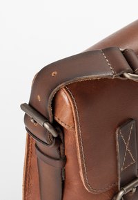 Angolo di una borsa in pelle marrone con una robusta tracolla in metallo, cuciture visibili e una superficie liscia e strutturata.