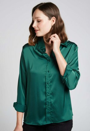 Blouse en satin vert avec col, fermeture à boutons et manches retroussées. Présentant une texture à pois subtile pour un détail supplémentaire.