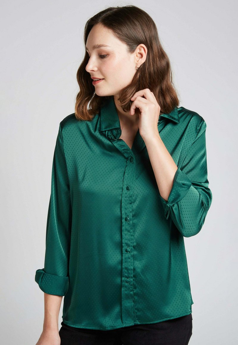 Blouse en satin vert avec col, fermeture à boutons et manches retroussées. Présentant une texture à pois subtile pour un détail supplémentaire.