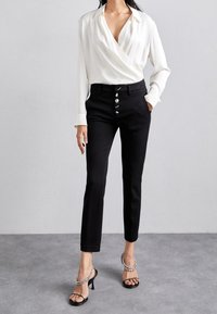 Weiße Wickelbluse mit langen Ärmeln, kombiniert mit hoch taillierten schwarzen Hosen mit silbernen Knopfdetails. Dazu schwarze Riemchenabsätze.