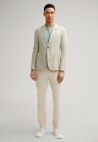 Hellgrauer, strukturierten Blazer mit zwei Vordertaschen, darunter ein mintgrünes Hemd, cremefarbene Hose und weiße Sneaker. Neutrale Farbpalette.
