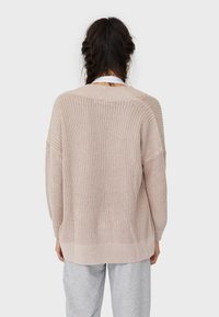 Ljusrosa stickad tröja med lös passform, nedre axlar och ribbad fåll. Strukturmönster ger visuell intresse. Bakvy visas.