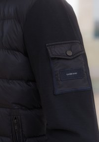 Jūras zila jaka ar kviltotu pūkainu audumu un teksturētām adītām piedurknēm. Iekļauj pogājamu kabatu ar etiķeti, kas lasa "GABBIANO."