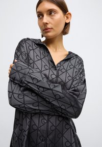 Cămașă gri cu mâneci lungi, cu un model abstract negru, având guler, manșete cu nasturi și croială lejeră. Textură netedă.