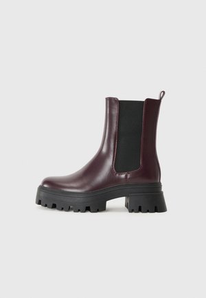 Bota de tornozelo de couro burgundy com painéis laterais elásticos, sola grossa de borracha preta e uma aba na parte de trás. Apresenta uma textura suave e salto bloco.