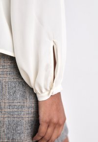 Krēmkrāsas blūze ar puffed piedurknēm, kurai ir pogu detaļa pie aproces un izdaiļojums pie plaukstas. Apvienota ar rūtainu svārku.