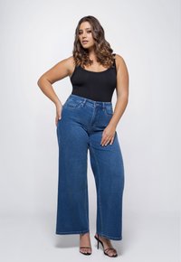 Blauwe wijde jeans met een hoge taille, gemaakt van denim, gecombineerd met een zwarte tanktop. Model staat tegen een effen achtergrond.