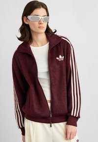 Pessoa a usar uma jaqueta zip-up oversized marrom da Adidas, uma t-shirt cropped branca, calças creme e óculos de sol futuristas prateados, contra um fundo simples.
