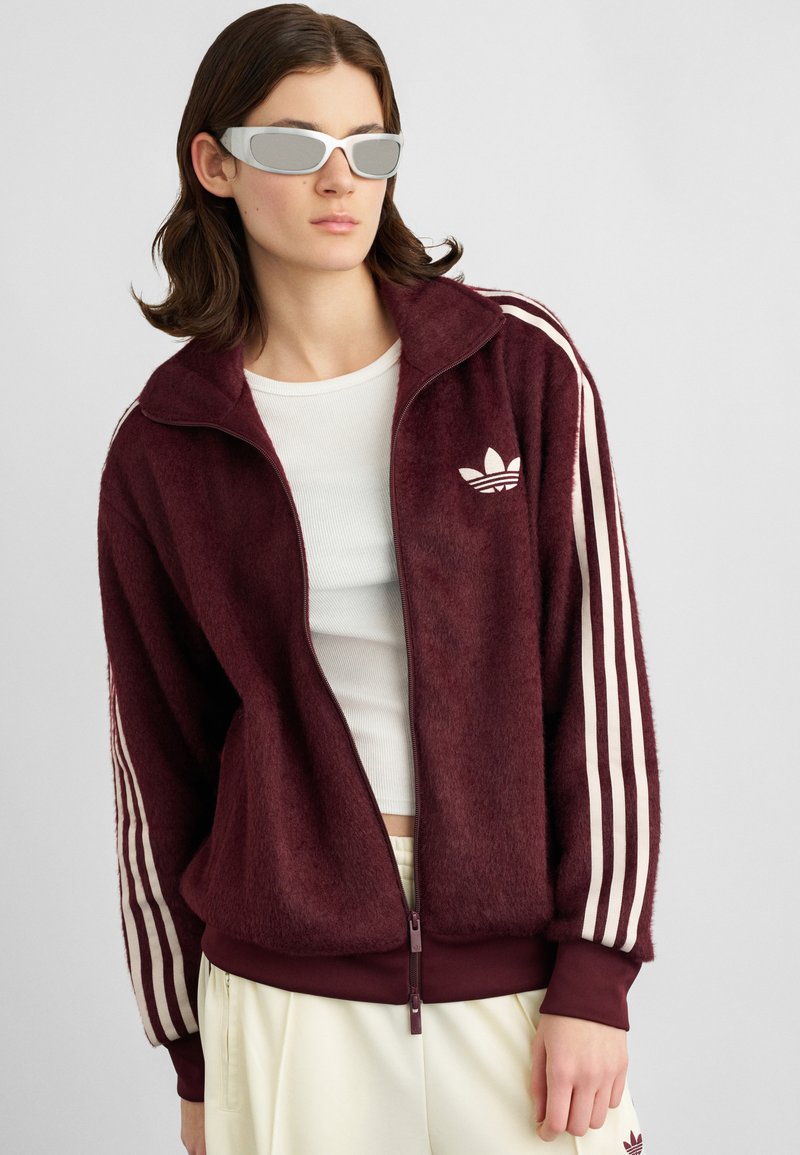 Pessoa a usar uma jaqueta zip-up oversized marrom da Adidas, uma t-shirt cropped branca, calças creme e óculos de sol futuristas prateados, contra um fundo simples.