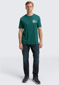 Camiseta de algodón verde con cuello redondo y mangas cortas, con texto gráfico blanco en el lado izquierdo del pecho. Lleva puesta con vaqueros oscuros y zapatillas.