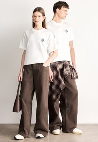 Fehér rövid ujjú pólók fekete gyémántmintával, barna bő szárú nadrággal párosítva; derékban megkötött kockás ing.